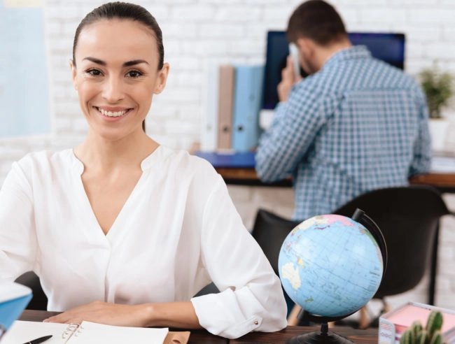 The travel agent keeps tickets for the plane in the travel agency. She offers them to clients. She smiles. On the table she has a toy plane, a laptop.